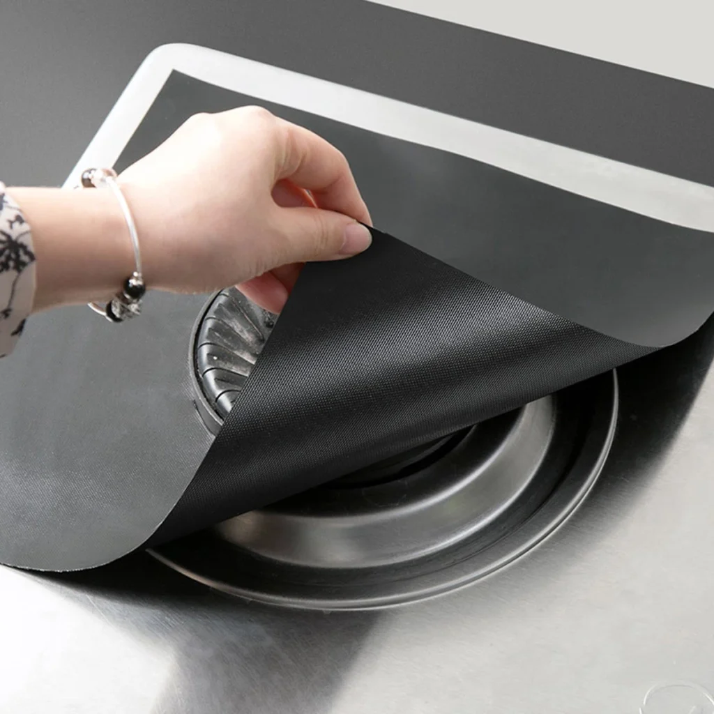 A hand placing a 0.2mm heat-resistant PTFE gas stove liner onto a stainless steel stovetop, demonstrating perfect fit and easy installation.