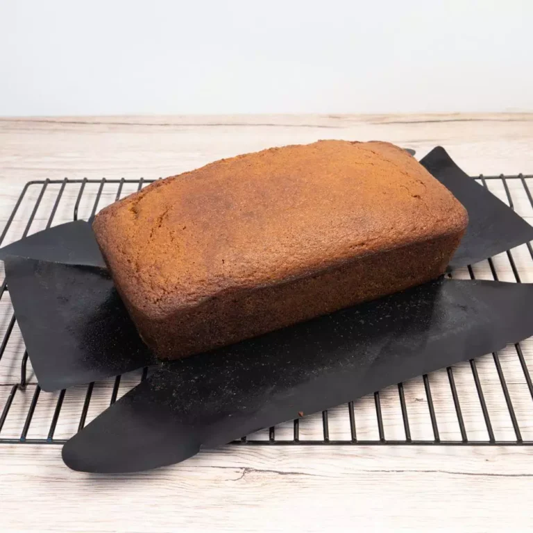 A baked pound bread block being lifted out seamlessly using a black non-stick PTFE loaf tin liner sheet on a cooling rack for bulk B2B wholesale.