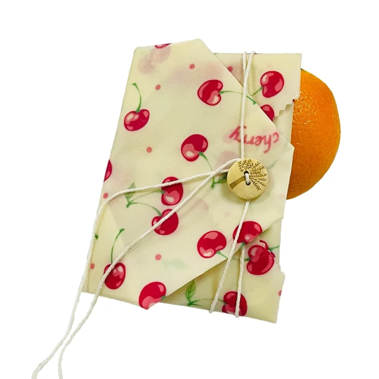 A premium single-piece organic **beeswax wrap with button factory** cotton sheet, wrapped around a fresh orange with an engraved wooden button and cotton string.
